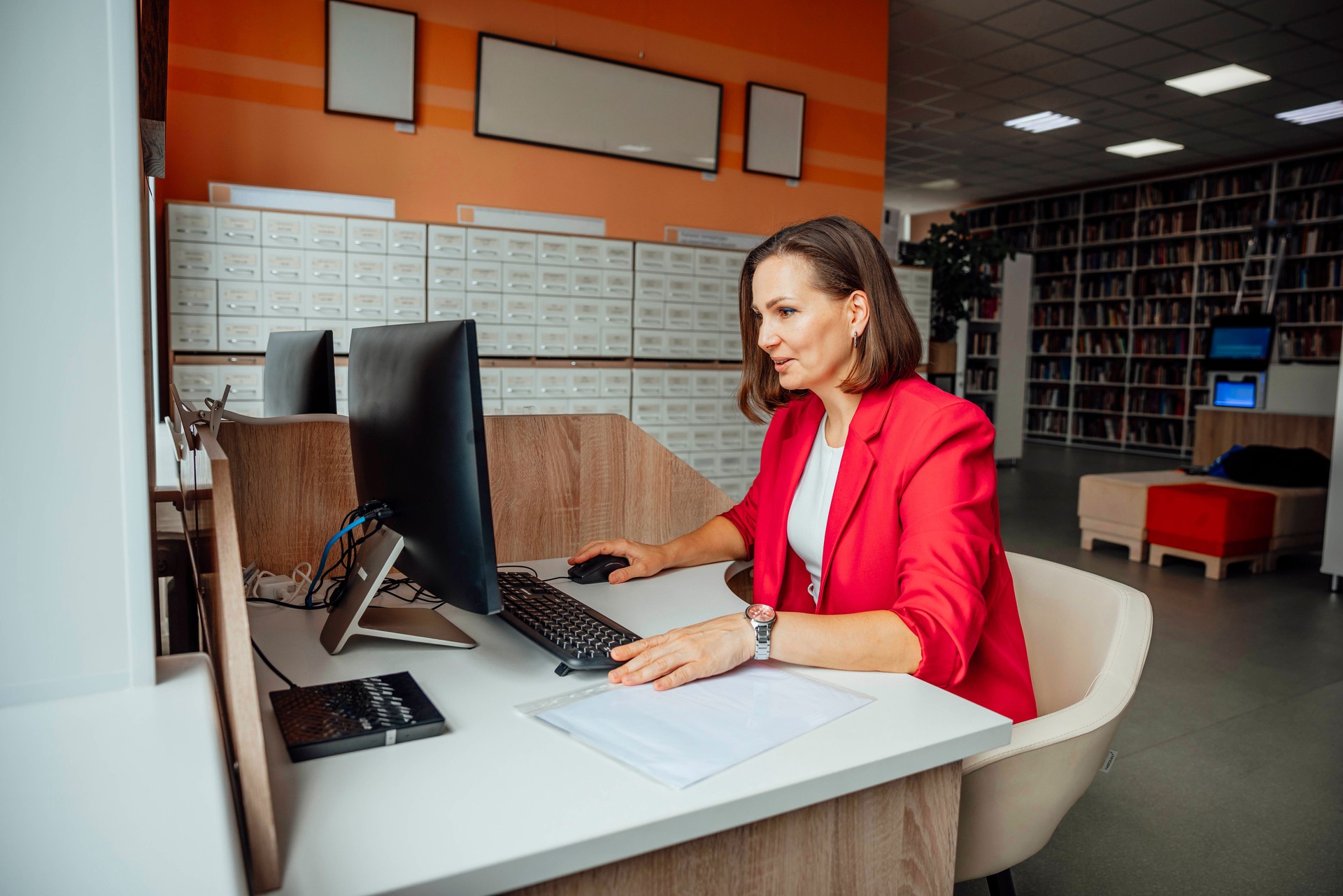 A young Caucasian woman with brown hair works at a computer in a modern coworking office. The space features wooden furniture and a library in the background. A young Caucasian woman with brown hair works at a computer in a modern coworking office. The space features wooden furniture and a library in the background.